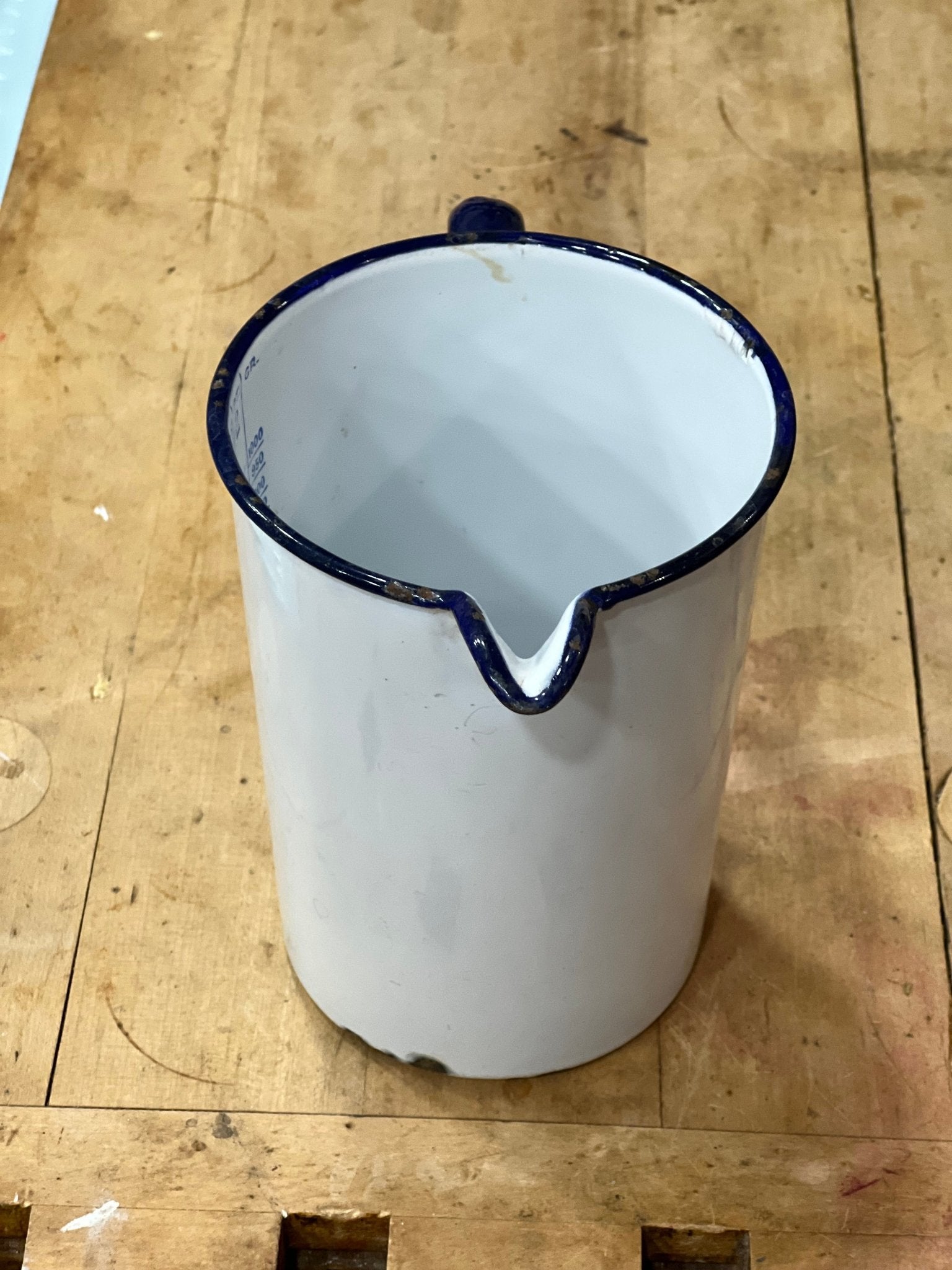 A white enamel pitcher with a dark blue rim and handle, shown from a top-down view on a wooden workbench.