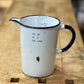 A vintage white enamel measuring pitcher with a dark blue rim, handle, and printed measurement markings, resting on a rustic wooden workbench.
