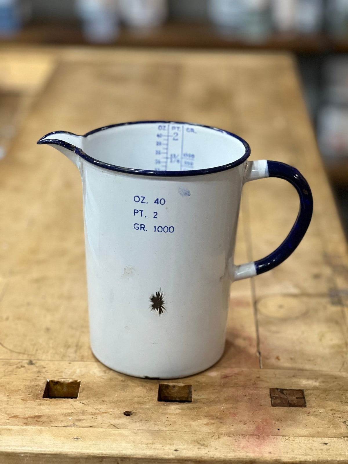 A vintage white enamel measuring pitcher with a dark blue rim, handle, and printed measurement markings, resting on a rustic wooden workbench.