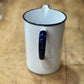 A white enamel pitcher with a dark blue rim and handle, featuring a small gold accent on the handle, set on a rustic wooden surface.