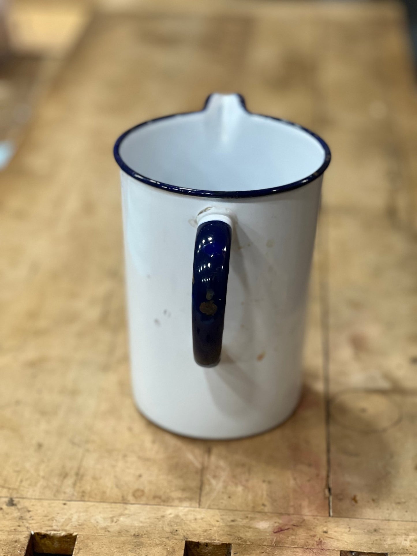 A white enamel pitcher with a dark blue rim and handle, featuring a small gold accent on the handle, set on a rustic wooden surface.