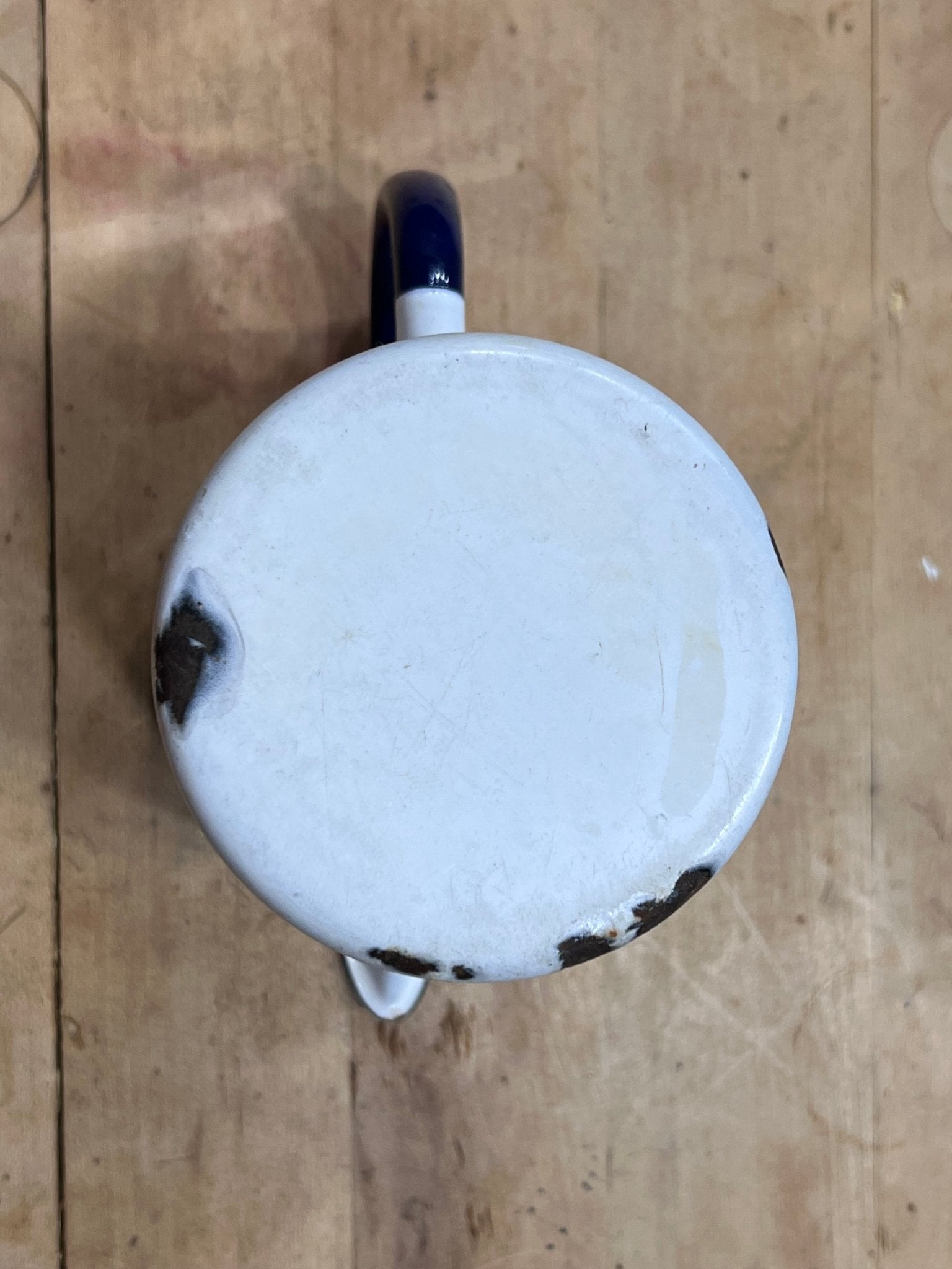 A top-down view of a vintage white enamel mug with a dark blue handle, resting on a light brown wooden surface.