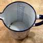 A vintage enamel pitcher viewed from above, featuring a white body with a dark blue rim, handle, and printed measurement markings inside.
