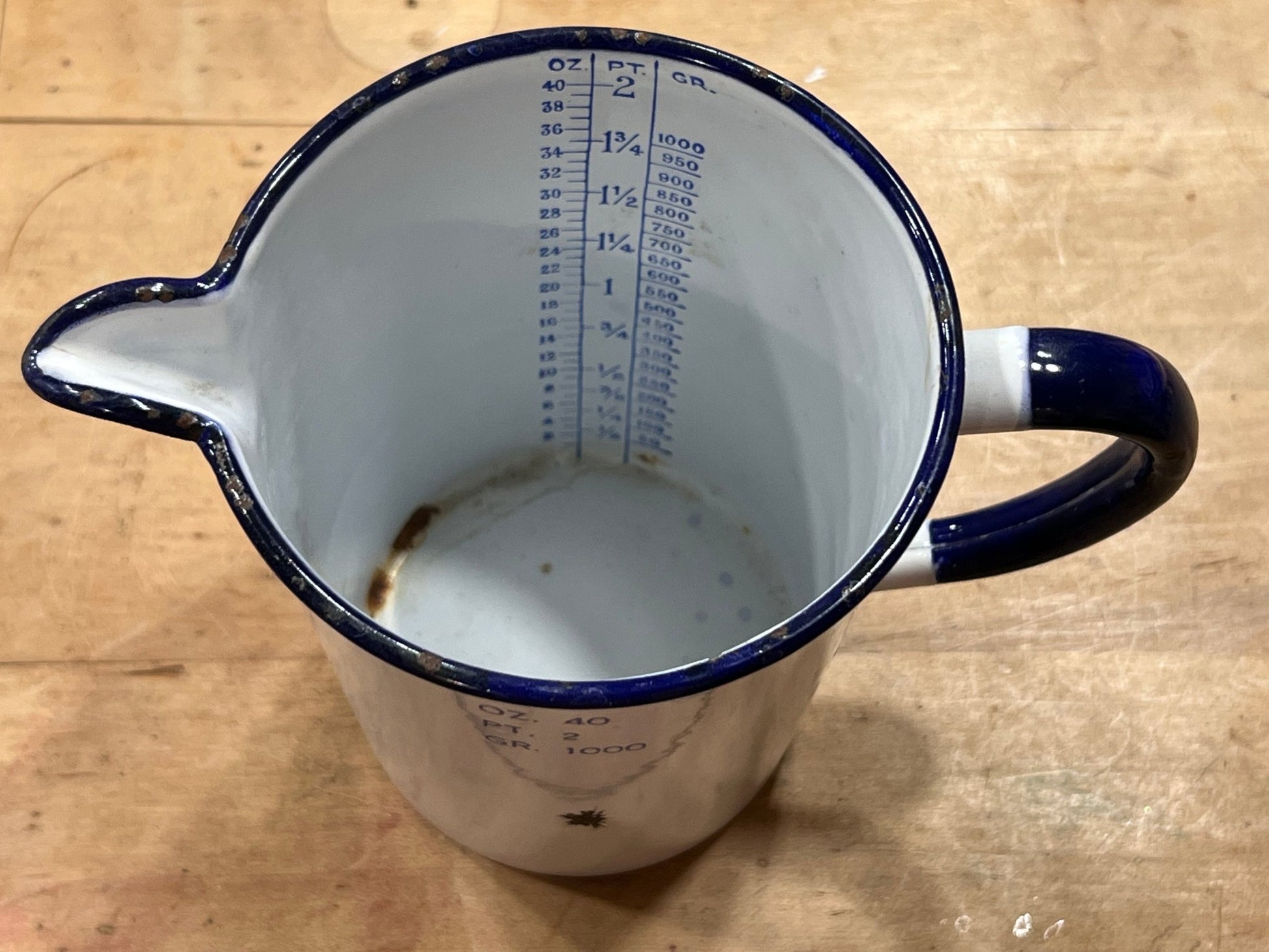 A vintage enamel pitcher viewed from above, featuring a white body with a dark blue rim, handle, and printed measurement markings inside.