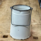 A vintage white enamel bucket with a black metal rim and wire handle, resting on a rustic wooden workbench.