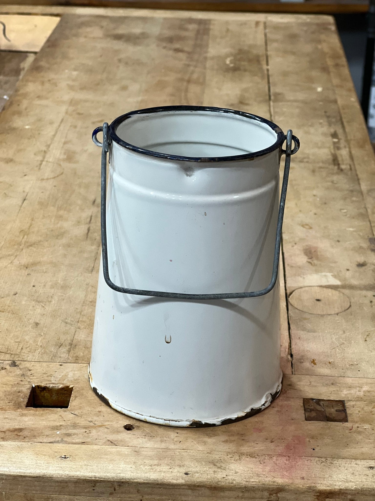 A vintage white enamel bucket with a black metal rim and wire handle, resting on a rustic wooden workbench.