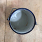 A top-down view of a vintage enamel paint can with a white interior, dark blue rim, and metal handle, resting on a light-colored wooden plank surface.