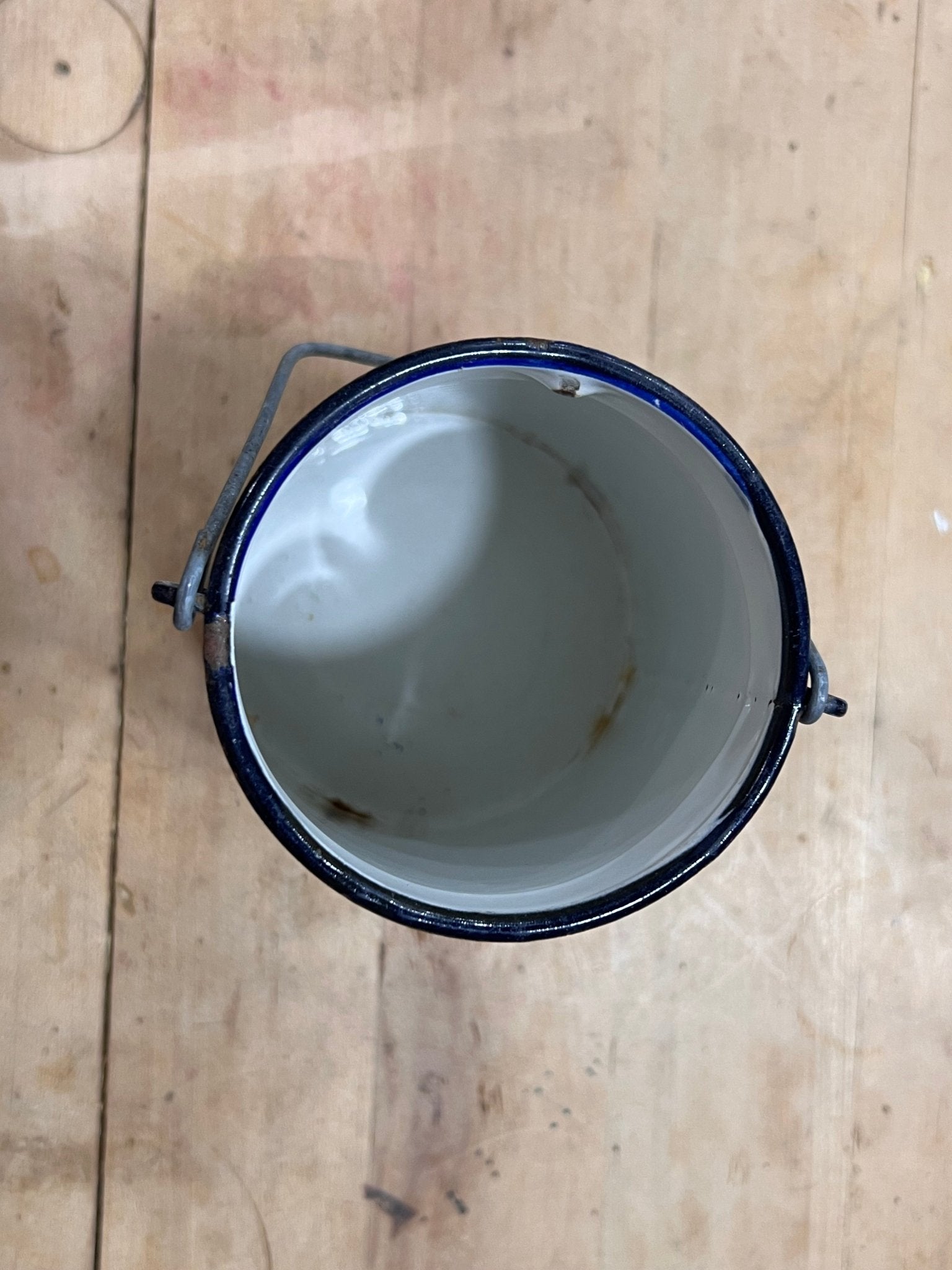 A top-down view of a vintage enamel paint can with a white interior, dark blue rim, and metal handle, resting on a light-colored wooden plank surface.