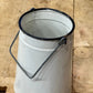 A white enamel paint can with a black rim and a metal loop handle, sitting on a rustic wooden plank surface.