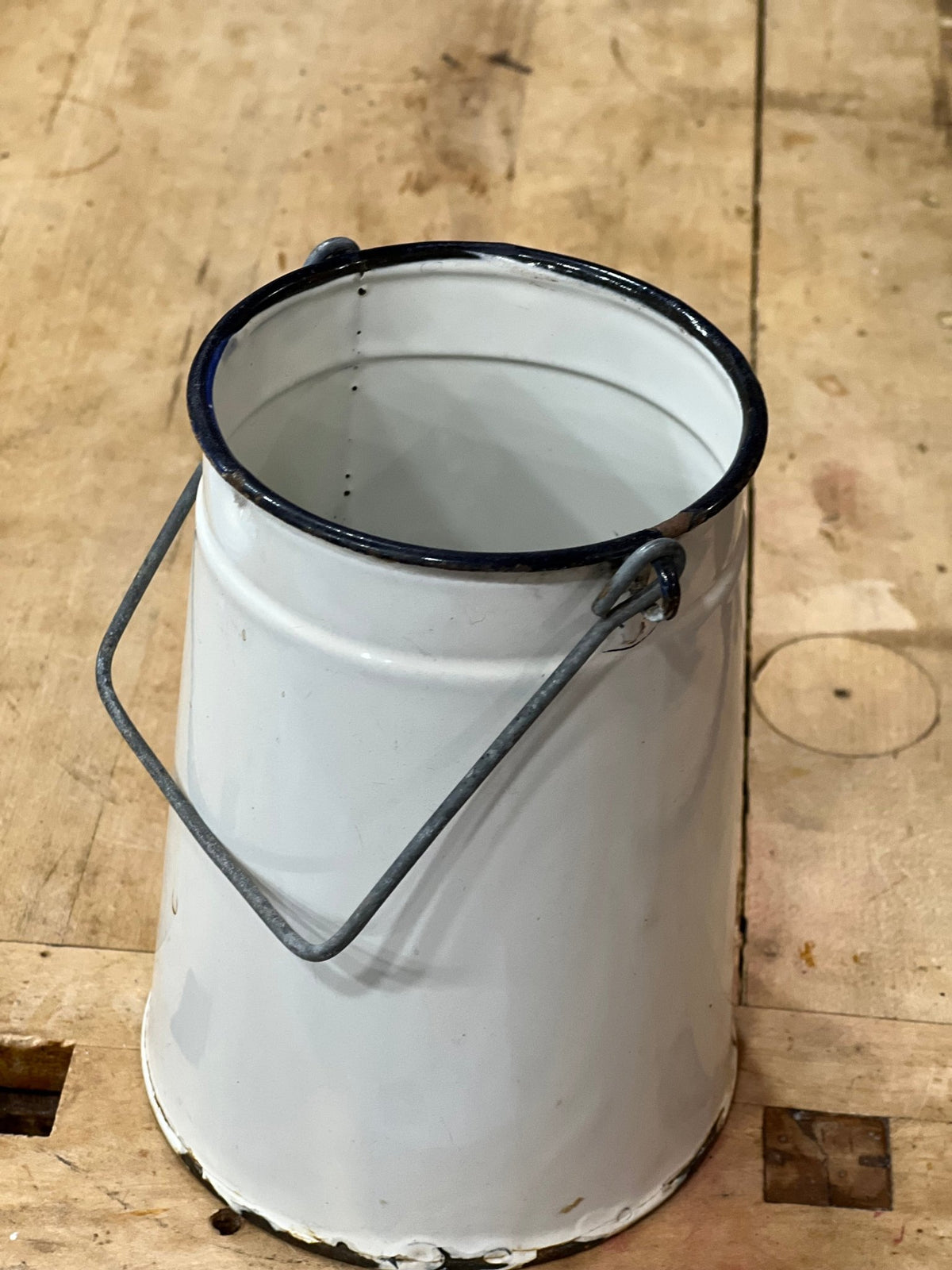 A white enamel paint can with a black rim and a metal loop handle, sitting on a rustic wooden plank surface.