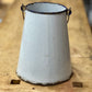 A vintage white enamel bucket with a conical shape, featuring dark metal handles and a weathered, distressed finish, resting on a wooden workbench.