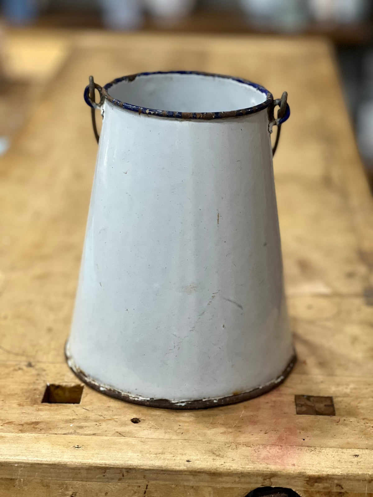 A vintage white enamel bucket with a conical shape, featuring dark metal handles and a weathered, distressed finish, resting on a wooden workbench.