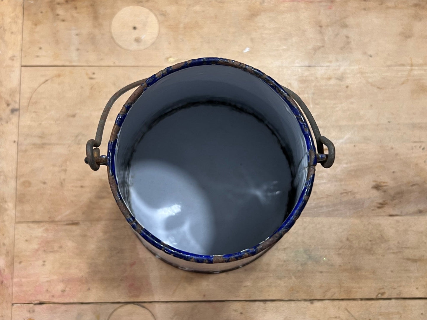 An open metal paint can filled with dark gray paint, viewed from above on a light wood plank surface.