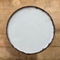 A round white enamel or plastic lid with a dark brown rim, viewed from directly above on a light brown wooden surface.