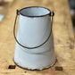 A vintage white enamel bucket with a dark metal wire handle, resting on a rustic wooden workbench.