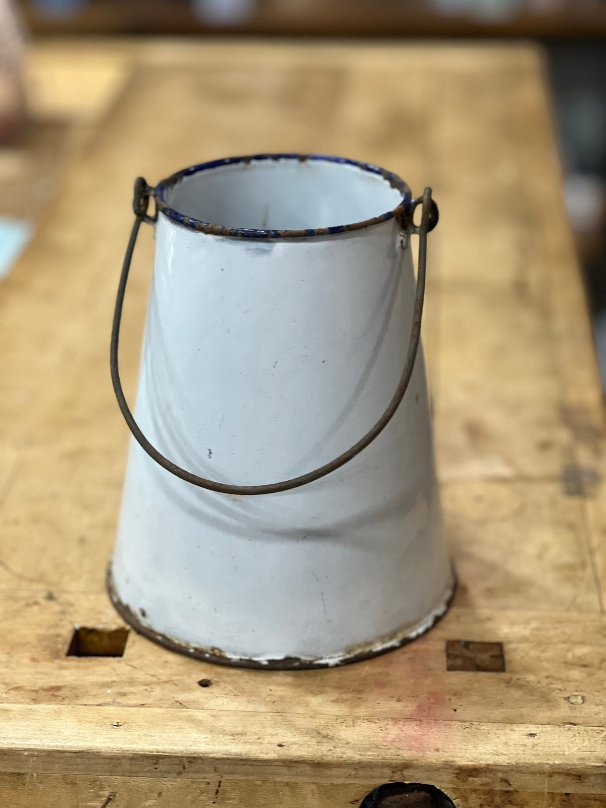 A vintage white enamel bucket with a dark metal wire handle, resting on a rustic wooden workbench.
