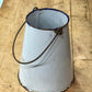 A vintage white enamel bucket with a blue rim and a dark metal wire handle, resting on a wooden workbench.