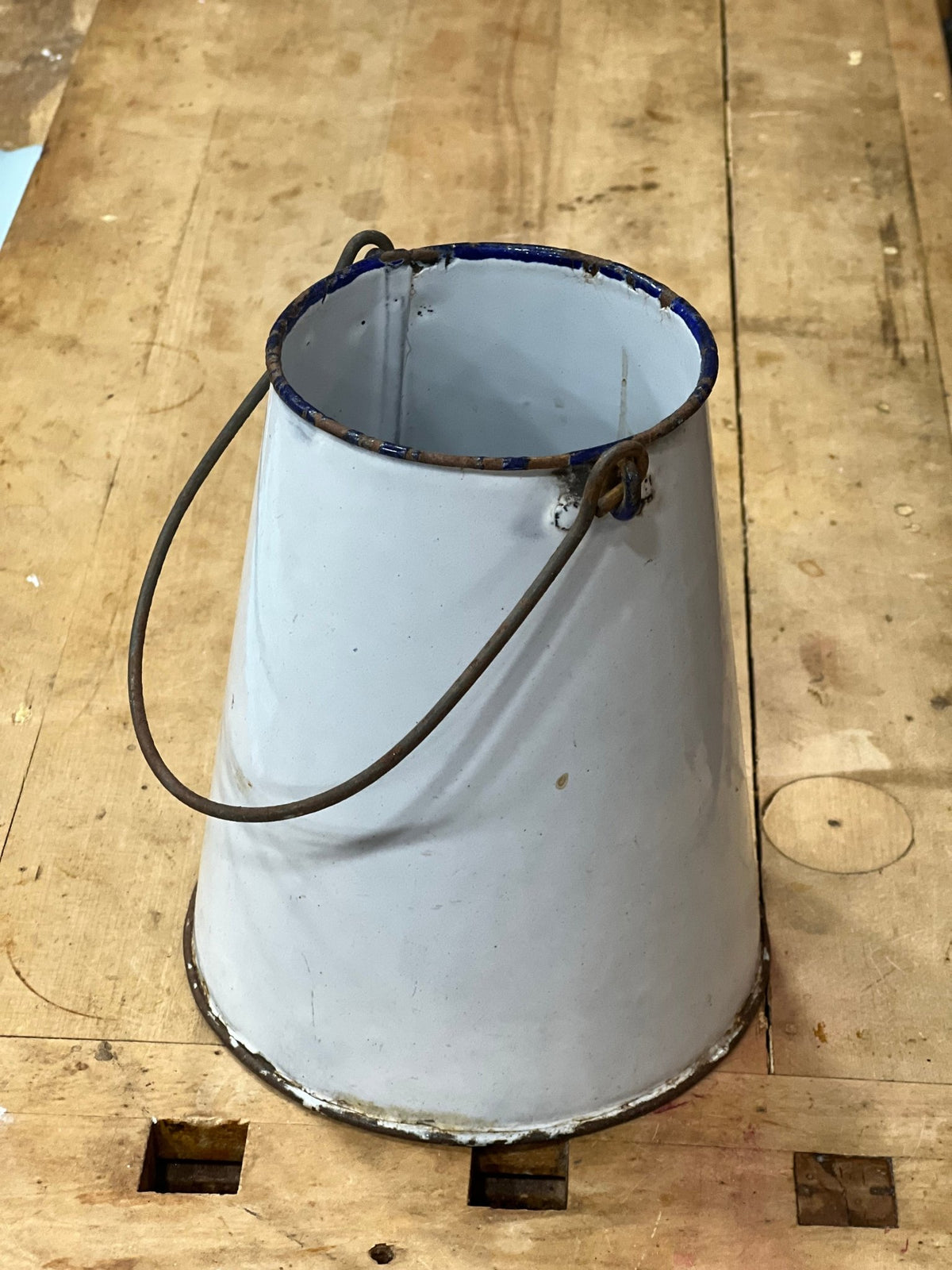 A vintage white enamel bucket with a blue rim and a dark metal wire handle, resting on a wooden workbench.
