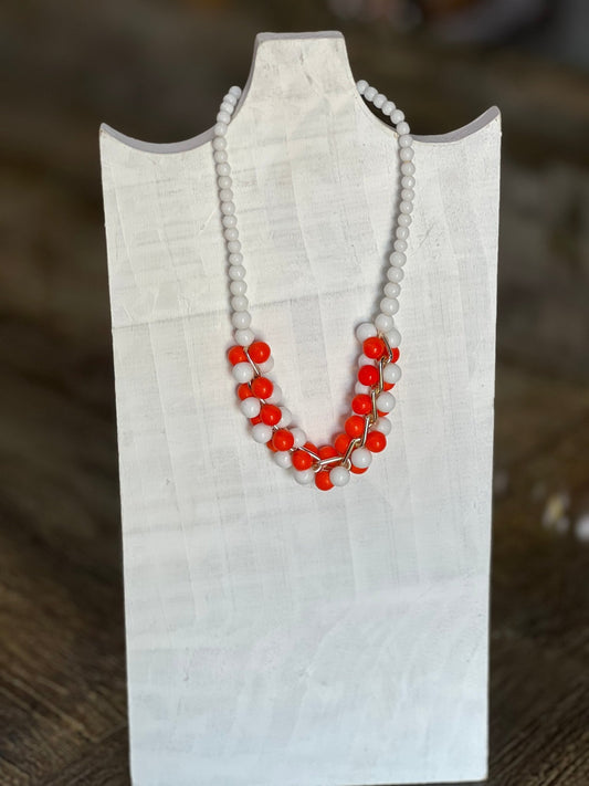 A beaded necklace with white round beads and bright red-orange bead clusters, displayed on a white fabric jewelry bust against a blurred brown background.